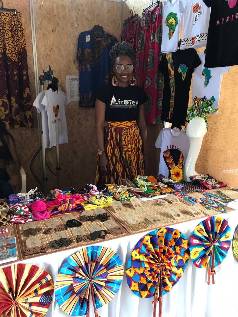Emprendedora de Afrotex sonriendo en su stand de la feria, rodeada de ropa con estampados africanos, turbantes, aretes de madera y abanicos de colores
