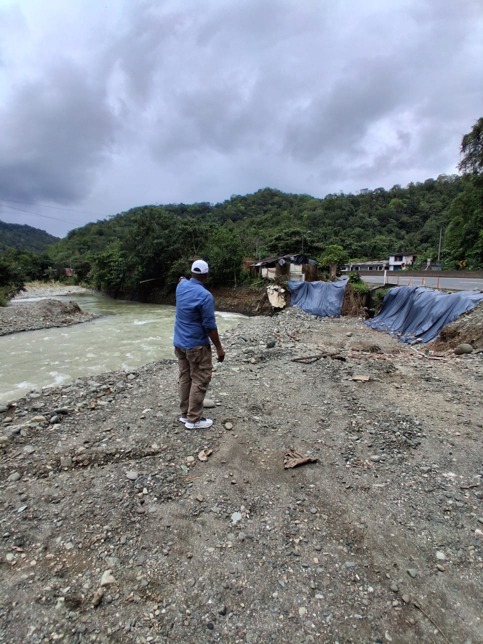 Un hombre con camisa azul y gorra blanca señala la ribera del río Dagua, donde hay viviendas cubiertas con lonas junto a la carretera